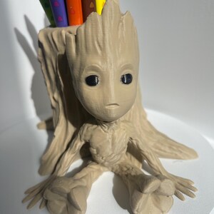 Puede incluir: Un portalápices de color beige con la forma del personaje Groot de Guardianes de la Galaxia. El soporte tiene un diseño detallado y texturizado y contiene varios bolígrafos de colores. El portalápices está sobre una superficie blanca.