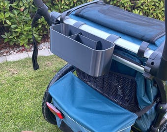 Può includere: Un organizer per passeggino grigio scuro con più scomparti è attaccato a un passeggino blu e nero. Il passeggino ha un manico nero e un vano portaoggetti blu. L'immagine mostra anche il testo "Happy Druck" in un cerchio rosa.
