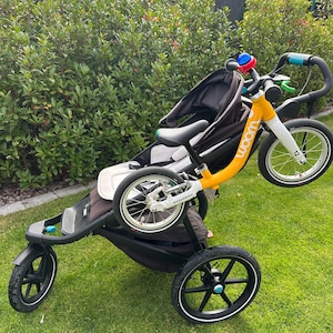 Könnte beinhalten: Ein gelb-weißes Laufrad mit schwarzen Reifen ist an einem schwarzen Kinderwagen befestigt. Auf dem Fahrrad ist das Wort "woom" auf dem Rahmen aufgedruckt. Der Kinderwagen hat einen schwarzen Sitz und einen grauen Rahmen mit drei schwarzen Rädern auf einem grünen Rasen.