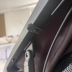 Può includere: Primo piano di un telaio di passeggino grigio scuro con interno in tessuto nero. L'immagine evidenzia la cerniera del passeggino e un piccolo pezzo di plastica nera fissato con una vite. È visibile una tasca a rete nera.