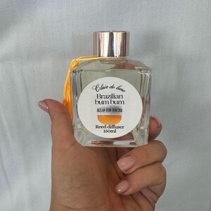 May include: A clear glass reed diffuser bottle with a gold cap and a yellow tassel. The label reads "Clair de lune Brazilian bum bum" and "Reed diffuser 150ml". The bottle is held in a hand.