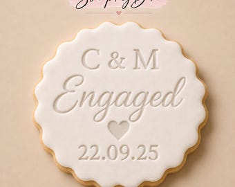 May include: A round, scalloped-edge cookie topped with smooth white icing. The text C & M, Engaged, and the date 22.09.25 are embossed into the icing, along with a small heart icon. The Stamp My Bake logo is visible at the top.