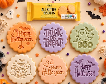 May include: A set of six round cookies with Halloween-themed embossed designs including ghosts, bats, pumpkins, and text reading Happy Halloween, Trick or Treat, and Boo, arranged below a yellow M&S Food All Butter Biscuits package.