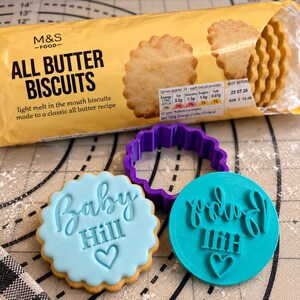 Puede incluir: Un paquete de galletas M&S Food All Butter, un cortador de galletas morado con bordes ondulados, una galleta glaseada azul claro con el texto Baby Hill y un coraz&oacute;n, y un sello de pl&aacute;stico turquesa con el texto Baby Hill y un coraz&oacute;n.