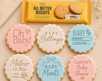 May include: Six round scalloped cookies with pastel icing and embossed text including Oh Baby, Baby Billings, Baby Loading, Mother to Bee, Baby Monti, and Lauren's Baby Shower, displayed with a yellow M&S All Butter Biscuits package.