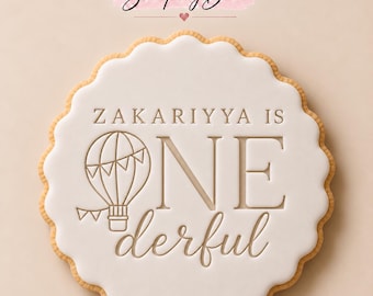 May include: A round scalloped cookie with white icing featuring the embossed text ZAKARIYYA IS ONE derful and a line art illustration of a hot air balloon. The Stamp My Bake logo is visible at the top of the image above the cookie.