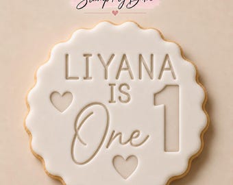 May include: A round scalloped cookie with white icing featuring the embossed text "LIYANA IS One" and the number "1". Two small heart shapes are embossed on the surface. The brand name "Stamp My Bake" appears above the cookie.