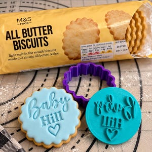 May include: A package of M&S Food All Butter Biscuits, a purple scalloped cookie cutter, a light blue iced cookie with the text Baby Hill and a heart, and a teal plastic stamp with the text Baby Hill and a heart embossed on it.