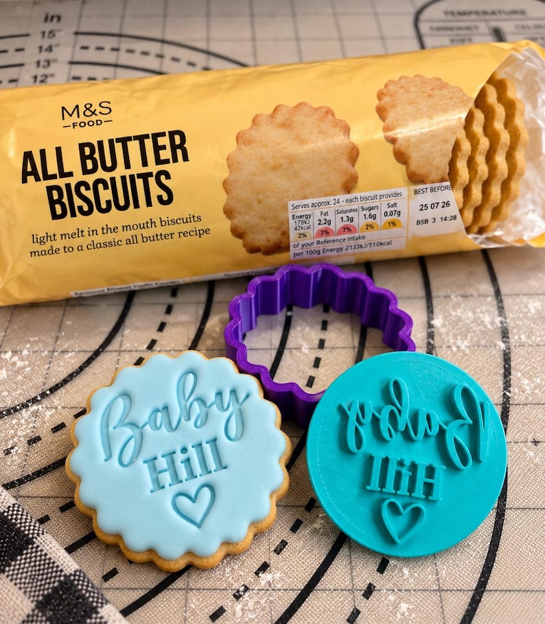 May include: A package of M&S Food All Butter Biscuits, a purple scalloped cookie cutter, a light blue iced cookie with the text Baby Hill and a heart, and a teal plastic stamp with the text Baby Hill and a heart embossed on it.