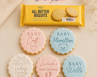 May include: Five round scalloped cookies with embossed text and designs, including baby names and shower themes, displayed alongside a yellow M&S All Butter Biscuits package. The cookies feature pink, blue, and white icing with various baby-themed motifs.