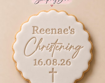 May include: A round, scalloped-edge cookie topped with smooth white icing. The icing is embossed with the text 'Reenae's Christening', the date '16.08.26', and a small cross symbol. The brand name 'Stamp My Bake' appears above the cookie.