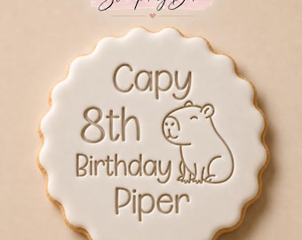 May include: A round scalloped cookie with white icing featuring the embossed text Capy 8th Birthday Piper and a line drawing of a capybara. Above the cookie, the text Stamp My Bake is displayed on a light pink brush stroke background.