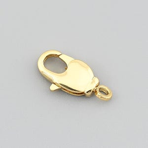 May include: A gold-colored lobster clasp, a jewelry component, is displayed against a gray background. The clasp is a shiny, metallic gold and has a curved shape with a spring-loaded mechanism for secure closure. It is designed for attaching to necklaces or bracelets.