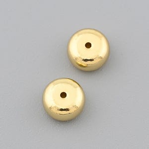 May include: Two small, round, gold-colored earrings with a polished finish. Each earring has a small, centered hole. The earrings are displayed against a plain, light gray background.