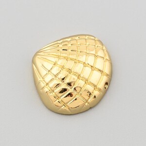 Op de afbeelding: Een goudkleurige, schelpvormige decoratieve knoop. De knoop heeft een gestructureerd, rasterachtig patroon over het oppervlak. De knoop staat tegen een effen, lichtgrijze achtergrond.