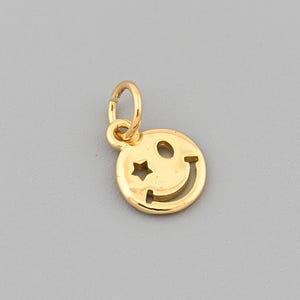 Op de afbeelding: Een goudkleurige bedel in de vorm van een lachend gezicht met een stervormig oog. De bedel heeft een kleine lus aan de bovenkant om aan een ketting of armband te bevestigen. De achtergrond is effen grijs.