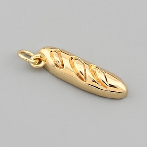 Könnte beinhalten: Ein goldfarbener Anhänger in Form eines Baguettes mit drei diagonalen Einschnitten. Der Anhänger hat eine kleine Schlaufe an einem Ende zum Befestigen an einer Kette oder einem Armband. Der Hintergrund ist einfarbig grau.