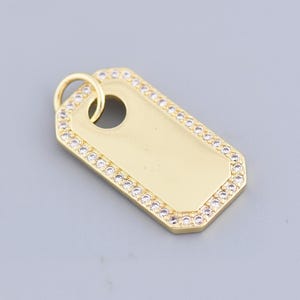 Puede incluir: Un colgante rectangular de color dorado con un pequeño agujero en la parte superior. El colgante está delineado con pequeños cristales transparentes. El fondo es de color gris claro liso. Este colgante es adecuado para collares.