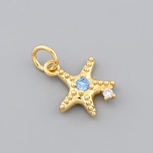 May include: A gold-colored starfish charm with a light blue gem in the center and a small white gem on one point. The charm has a textured surface and a small loop for attaching to a necklace or bracelet.
