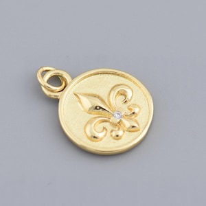 May include: A gold-colored pendant with a fleur-de-lis design. The pendant is circular with a textured surface and a small diamond accent in the center of the design. The pendant has a small loop for a chain.