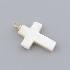 May include: A white cross pendant with a gold-colored bail. The cross is made of a pearlescent material and has a smooth, polished finish. The pendant is set against a gray background.