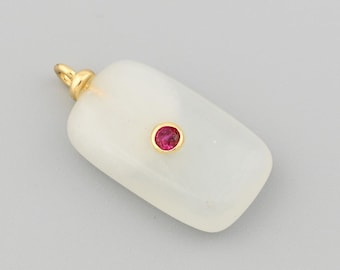 Puede incluir: Colgante rectangular con una fianza dorada y una pequeña gema roja redonda en el centro. El colgante es de color blanco cremoso y tiene una superficie lisa. La fianza es dorada.