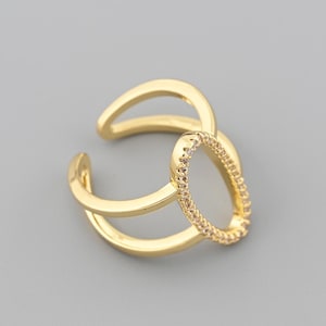 May include: A gold-colored ring with an open oval design, embellished with small, sparkling stones. The ring features a double-band design and is set against a gray background. This piece of jewelry is a stylish accessory.
