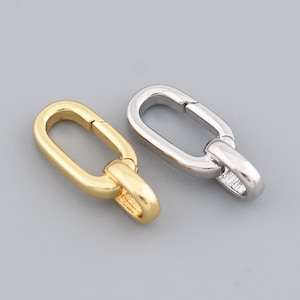 Pode incluir: Duas fivelas ovais, uma dourada e outra prateada, sobre uma superfície cinza. As fivelas são projetadas com um mecanismo de dobradiça para um fechamento seguro, ideais para a criação de joias ou projetos de artesanato.