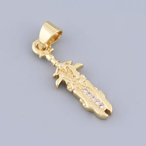 May include: A gold-colored pendant in the shape of a sword. The sword pendant has a decorative hilt and a blade adorned with small, clear gemstones. The pendant is suspended from a small loop for a chain.