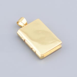 Könnte beinhalten: Ein rechteckiger goldener Anhänger mit einer kleinen Schlaufe oben. Der Anhänger hat eine glatte, reflektierende Oberfläche und ein Scharnierdesign. Der Anhänger ist wahrscheinlich aus Gold und vor einem grauen Hintergrund platziert.