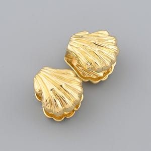 May include: Two gold-colored seashell-shaped earrings. The earrings have a detailed, ridged texture, mimicking the natural form of a seashell. The earrings are displayed against a plain gray background.