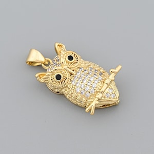 Könnte beinhalten: Ein goldfarbener Eulenanhänger mit detailliertem Design. Der Körper der Eule ist mit kleinen, klaren Kristallen bedeckt und hat schwarze Augen. Der Anhänger hält einen kleinen Ast und hat eine Schlaufe für eine Kette.