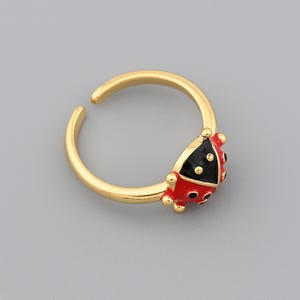 May include: A gold-colored adjustable ring featuring a ladybug design. The ladybug has a red body with black spots and a black head. The ring is set against a gray background.