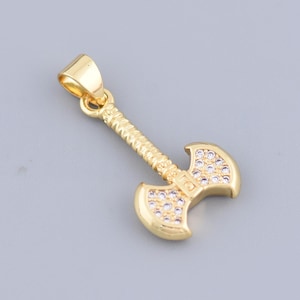 May include: A gold-colored pendant in the shape of a double-bladed axe. The axe head is adorned with small, clear gemstones. The handle has a textured design. The pendant is suspended from a small loop.