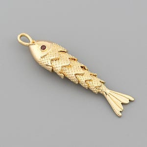 May include: A gold-colored fish pendant with a textured scale design and a small red gem for the eye. The fish has a loop at the top for hanging and a detailed tail. The pendant is set against a gray background.