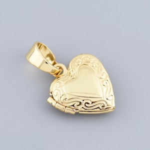 Op de afbeelding: Een goudkleurige hartvormige locket hanger. De locket heeft een sierlijk, gegraveerd ontwerp aan de voorkant en een kleine lus voor een ketting. De hanger wordt getoond tegen een lichtgrijze achtergrond.