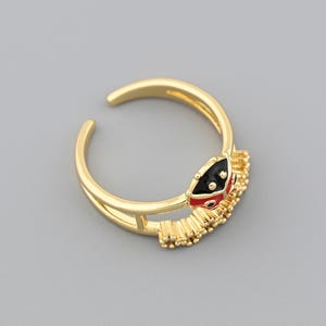 May include: A gold-colored adjustable ring with a snake design. The snake's head features black and red enamel detailing. The ring has a double-band design and is set against a gray background.