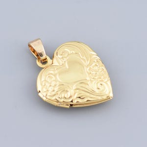 Op de afbeelding: Een goudkleurige hartvormige medaillonhanger met een sierlijk bloemmotief. De hanger heeft een kleine lus aan de bovenkant voor een ketting. Het medaillon is gesloten en het oppervlak is glad en reflecterend.