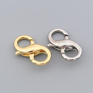 May include: Two metal lobster clasps, one gold-toned and one silver-toned, are displayed against a gray background. The clasps are in the shape of an 'S' with a spring-loaded lever for secure closure. These clasps are commonly used in jewelry making and crafting.