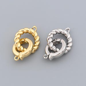 May include: Two metal jewelry clasps. One is gold-colored, the other is silver-colored. Both clasps have a twisted rope design around the circular clasp. Each clasp has a small loop and a hook for secure closure. The background is a solid gray.