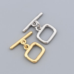 May include: Two toggle clasps, one silver-colored and one gold-colored, are displayed against a gray background. Each clasp consists of a rectangular loop and a bar. The gold clasp has a warm, shiny finish, while the silver clasp has a bright, metallic appearance.