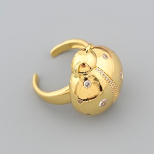 Könnte beinhalten: Ein goldfarbener Ring mit einem Marienkäfer-Design. Der Ring hat einen abgerundeten Körper mit kleinen, klaren Edelsteinen und einem strukturierten Band. Der Ring ist verstellbar und liegt auf grauem Hintergrund.