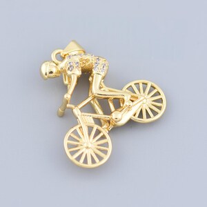 以下が含まれることがあります： 金色の自転車ペンダント。レーシングポジションのサイクリストが描かれています。ペンダントは、自転車のフレーム、ホイール、サイクリストの服装に複雑なディテールが施されています。サイクリストはヘルメットとショーツを着用しています。ペンダントは灰色の背景にあります。