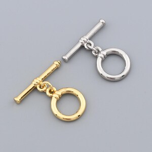 May include: Two toggle clasps, one gold-toned and one silver-toned, lie on a gray surface. Each clasp consists of a bar and a ring. The gold clasp is on the left, and the silver clasp is on the right.