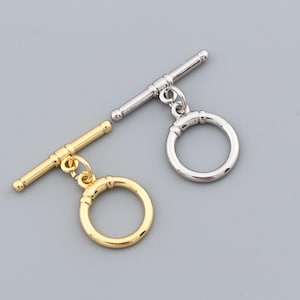 Pode incluir: Duas fechos de alternância, um dourado e outro prateado, contra um fundo cinzento. Cada fecho tem uma barra em T e um anel circular, para fazer joias. O fecho dourado está à esquerda, o prateado à direita.