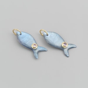 May include: Two fish-shaped pendants crafted from a smooth, iridescent blue material. Each pendant features a small, round, light pink gemstone set in a gold-coloured bezel. A small gold-coloured jump ring is attached to the top of each fish charm.