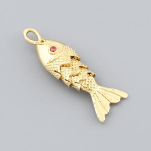 May include: A gold-colored fish pendant with a red gemstone eye. The pendant has a textured scale design and a loop at the top for hanging. The fish's tail is fanned out. The pendant is on a gray background.
