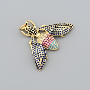 May include: A gold-colored bee-shaped brooch with blue, black, pink, and turquoise gemstones. The wings and body are adorned with small, round gemstones. The brooch has a detailed design and is set against a gray background.