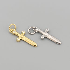 May include: Two small sword pendants. One is gold-coloured, the other is silver-coloured. Both have a loop at the top for hanging and are decorated with small, clear gemstones. The swords are displayed on a grey background.