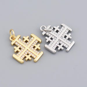 Może przedstawiać: Dwie zawieszki z krzyżem jerozolimskim. Jedna jest w kolorze złotym z przezroczystymi kamieniami, druga w kolorze srebrnym z przezroczystymi kamieniami. Obie mają pętelkę na łańcuszek. Krzyże są na szarym tle.